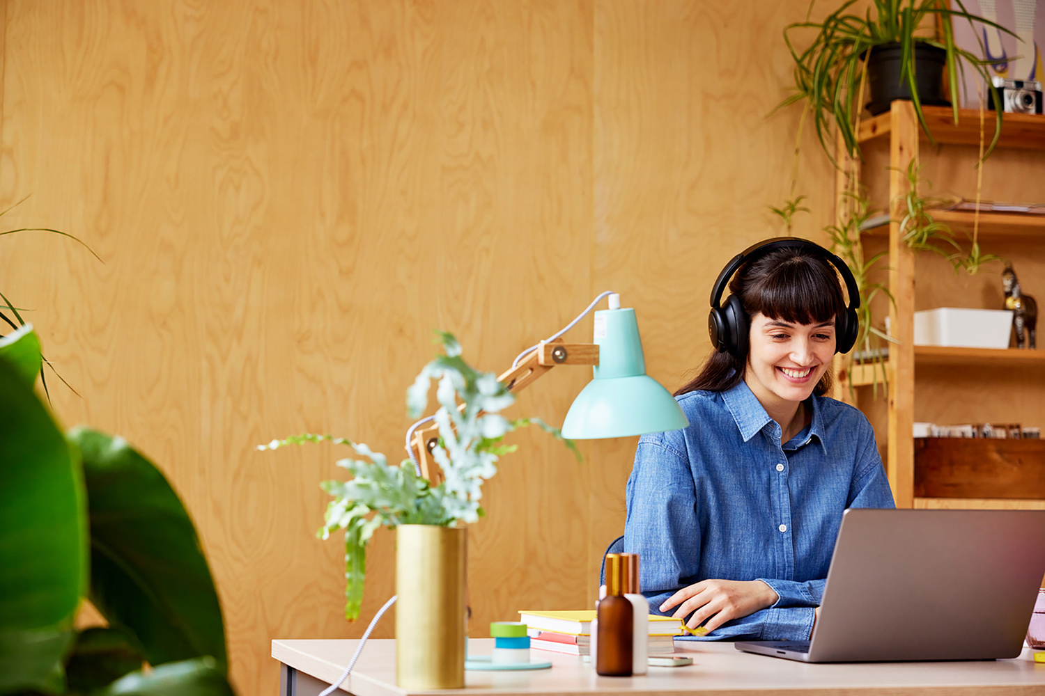 A person sitting at a desk, working on a laptop. The person has dark hair, bangs, and is wearing large black headphones and a blue button-down shirt. They appear to be smiling and looking at the laptop screen. The desk is light-coloured and has various items on it, including a light blue desk lamp, a gold vase with green plants, small containers, and a brown bottle. A laptop is open in front of the person, and their hands are positioned as if typing or using the trackpad. The background features a light brown wooden paneled wall and a wooden shelf unit holding plants and other decorative objects. The overall setting suggests a home office or a comfortable workspace where various online services, including Wifi and pure Fibre Internet, would be essential for remote work or communication.