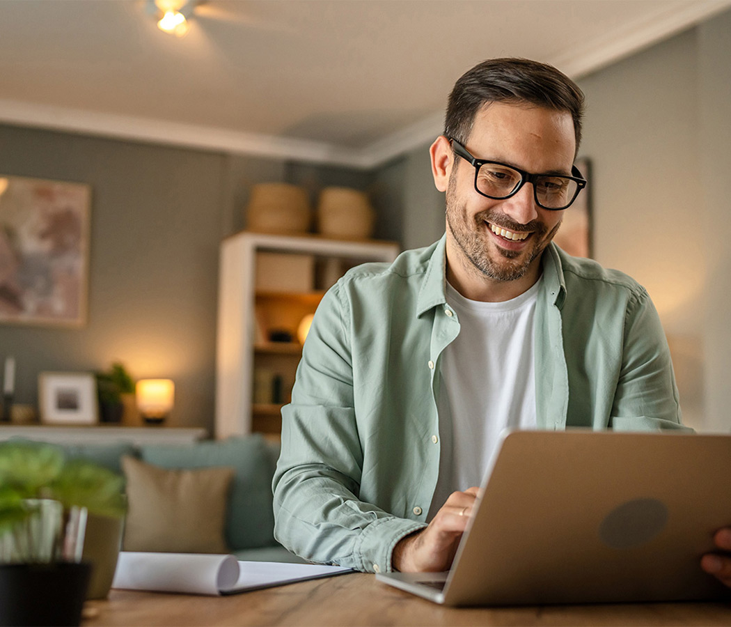Man in glasses using a laptop at home, connected to high-speed pure Fibre Internet, sitting at a wooden table with a notebook and pen. The room has soft lighting and modern decor.