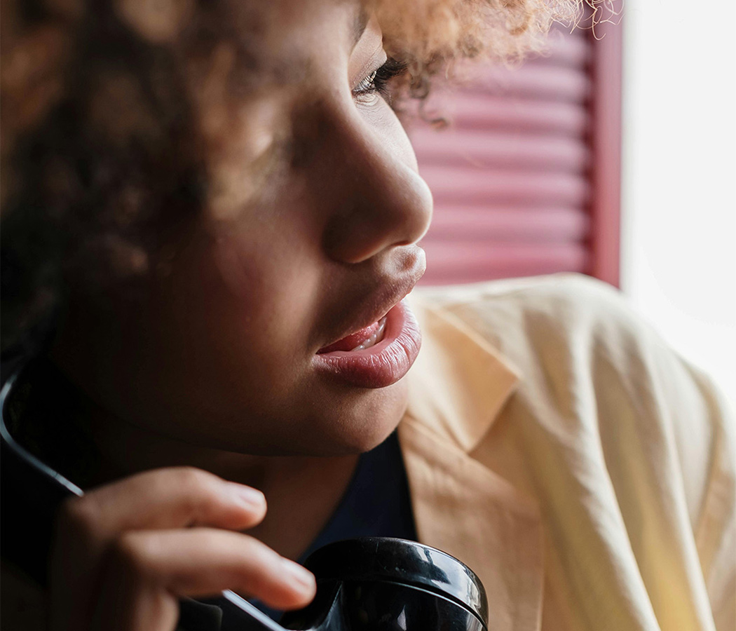 Close-up profile of a person with curly hair talking on a traditional black landline phone, with soft light coming through pink window blinds in the background. They appear engaged in conversation, highlighting the reliability of home phone services that many still value alongside modern technology. A dependable landline plan from a trusted provider ensures clear nationwide calling capability, offering an important communication option for households seeking comprehensive connectivity services.
