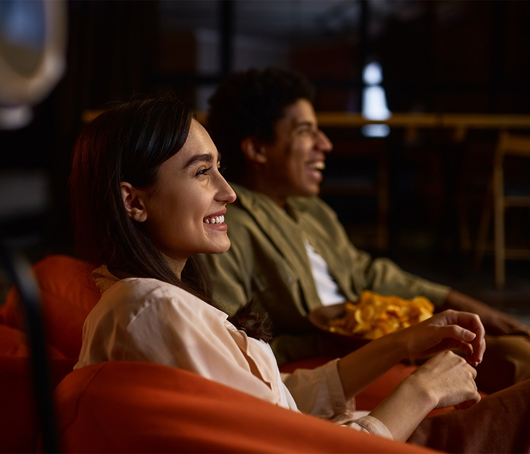 A man and woman enjoying entertainment at home, smiling as they sit on an orange couch in a dimly lit room. The woman in a light blouse and the man in a green shirt share a bowl of chips while watching something on TV. Their expressions of delight highlight how pure fibre internet plans deliver seamless streaming experiences, with high-speed wifi service ensuring buffer-free entertainment.
